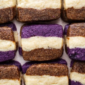 Rectangular ube coffee shortbread cookies with three distinct layers—purple on top, brown on the bottom, and a cream-colored center—are arranged neatly in rows on a flat surface.