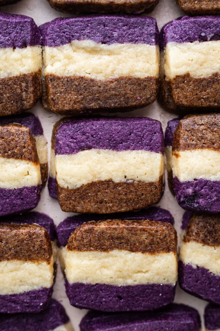 Rectangular ube coffee shortbread cookies with three distinct layers—purple on top, brown on the bottom, and a cream-colored center—are arranged neatly in rows on a flat surface.