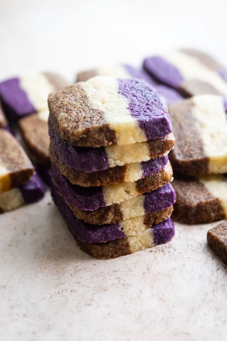 A stack of rectangular ube coffee shortbread cookies with three layers: brown, purple, and white. More cookies with the same pattern are scattered in the background on a light surface.