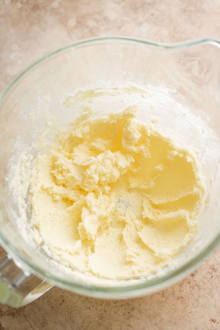 A glass mixing bowl filled with creamed butter and sugar, seen from above, on a light brown countertop.