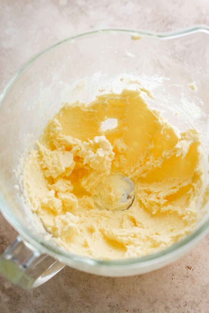A glass mixing bowl containing creamed butter and sugar on a light-colored countertop. The mixture appears pale and fluffy, with some scraped up the sides of the bowl.