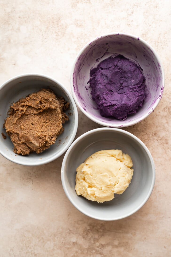 Three small bowls with different doughs: one brown, one yellow, and one purple, arranged in a triangle on a light, textured surface.