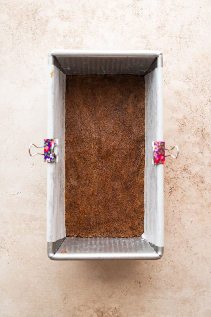 A rectangular metal loaf pan with a brown crust or baked layer at the bottom, lined with parchment paper held in place by two colorful binder clips, sits on a light beige surface.