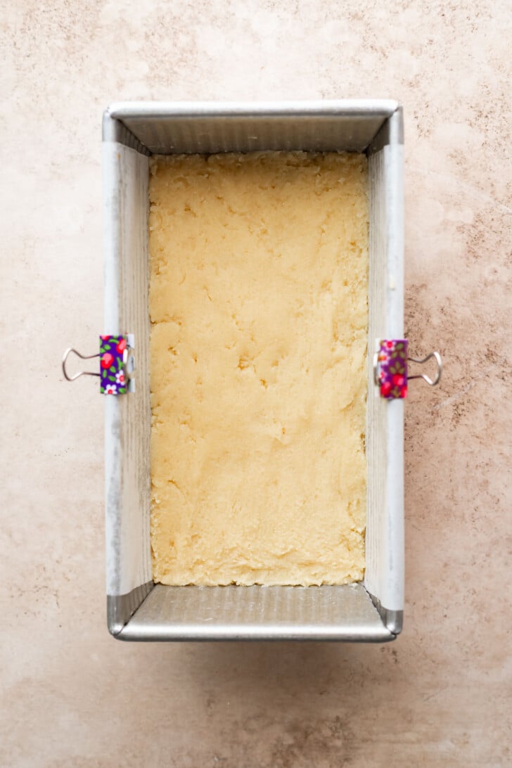 A loaf pan lined with parchment paper, held in place by two colorful binder clips, is filled with an even layer of raw dough or batter. The pan sits on a light, textured surface.
