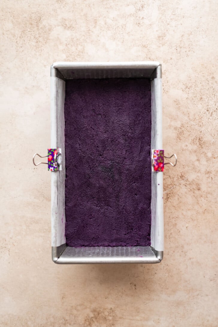 A rectangular metal loaf pan with a deep purple mixture inside, held with two colorful binder clips, sits on a light beige textured surface.