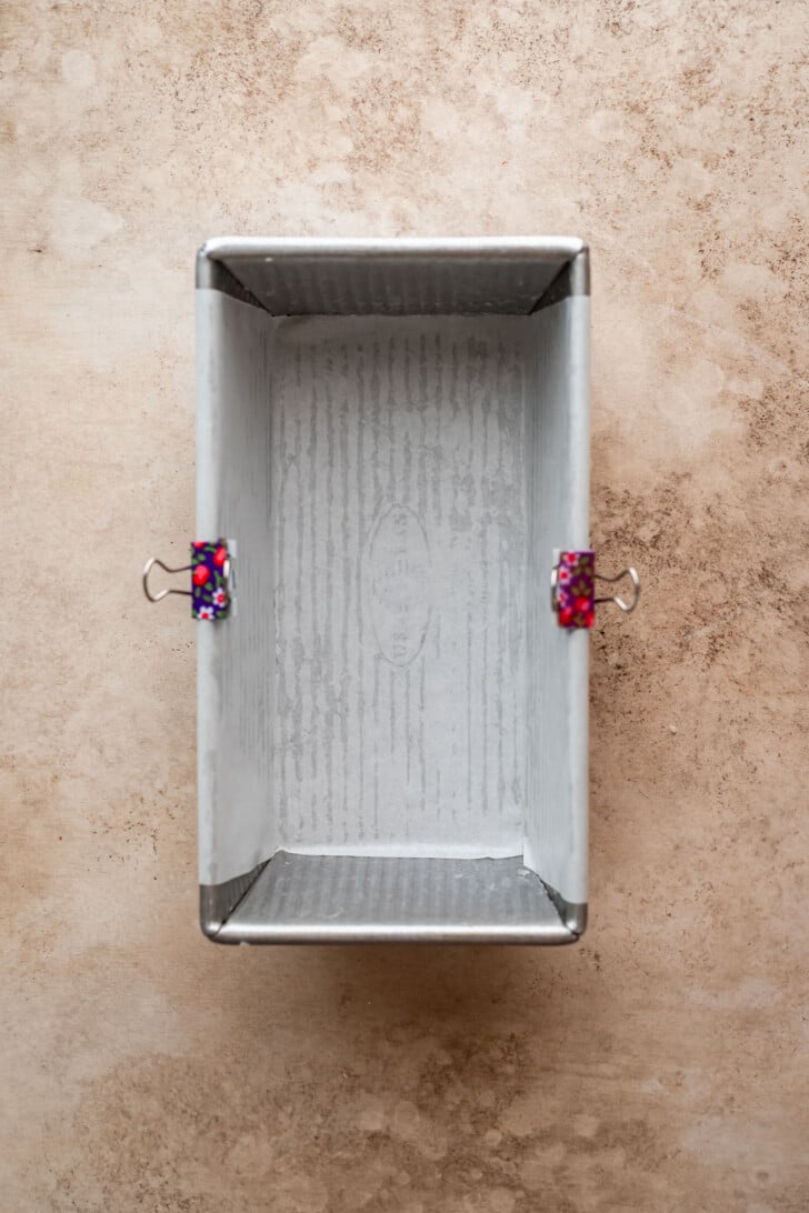 A rectangular metal loaf pan lined with parchment paper, held in place with two binder clips, sits on a light brown textured surface.