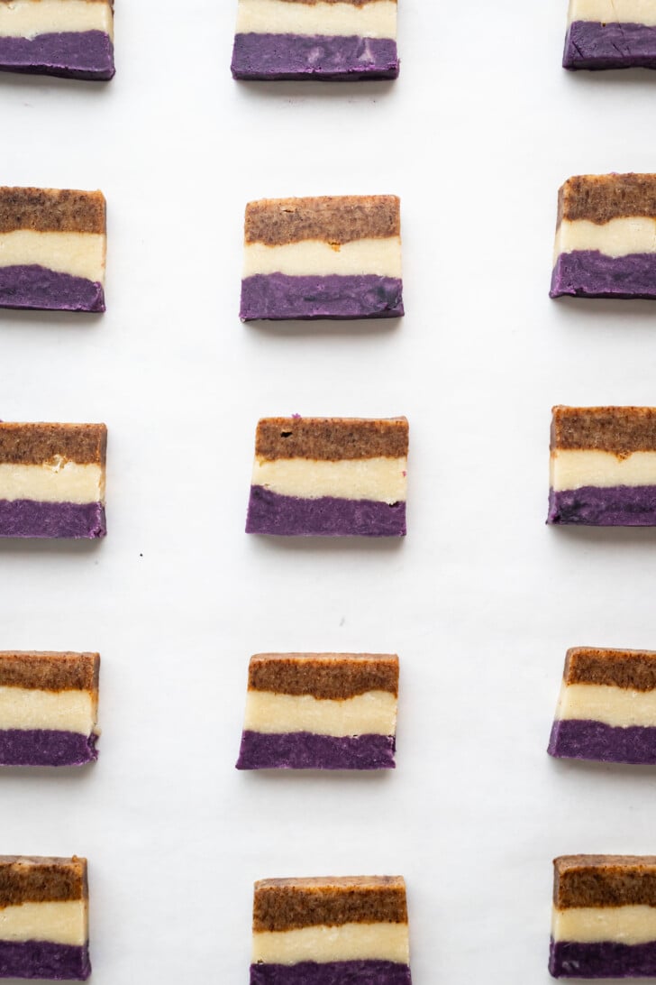 Rectangular layered desserts with purple, cream, and brown layers are neatly arranged in rows on a white background.
