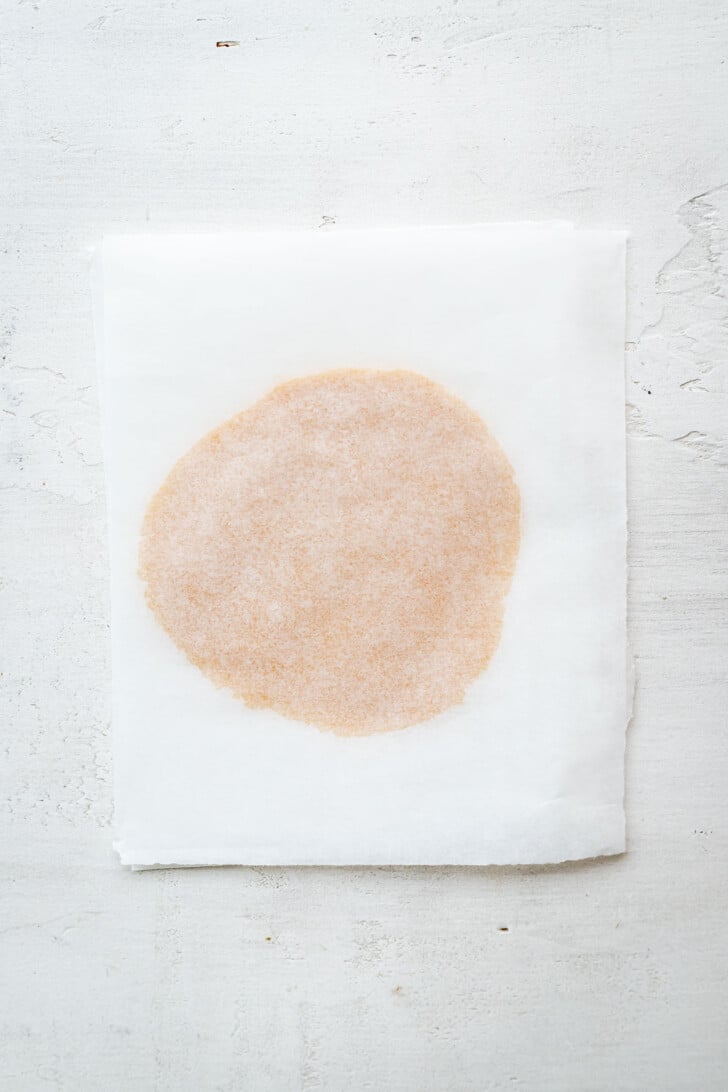 A circular light brown dough between 2 pieces of white parchment paper, placed on a textured white surface.