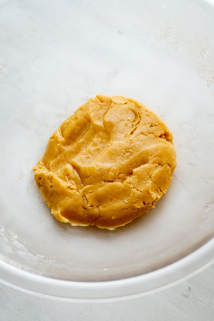 A round ball of yellow mooncake dough sits in the center of a clear glass mixing bowl on a light surface.