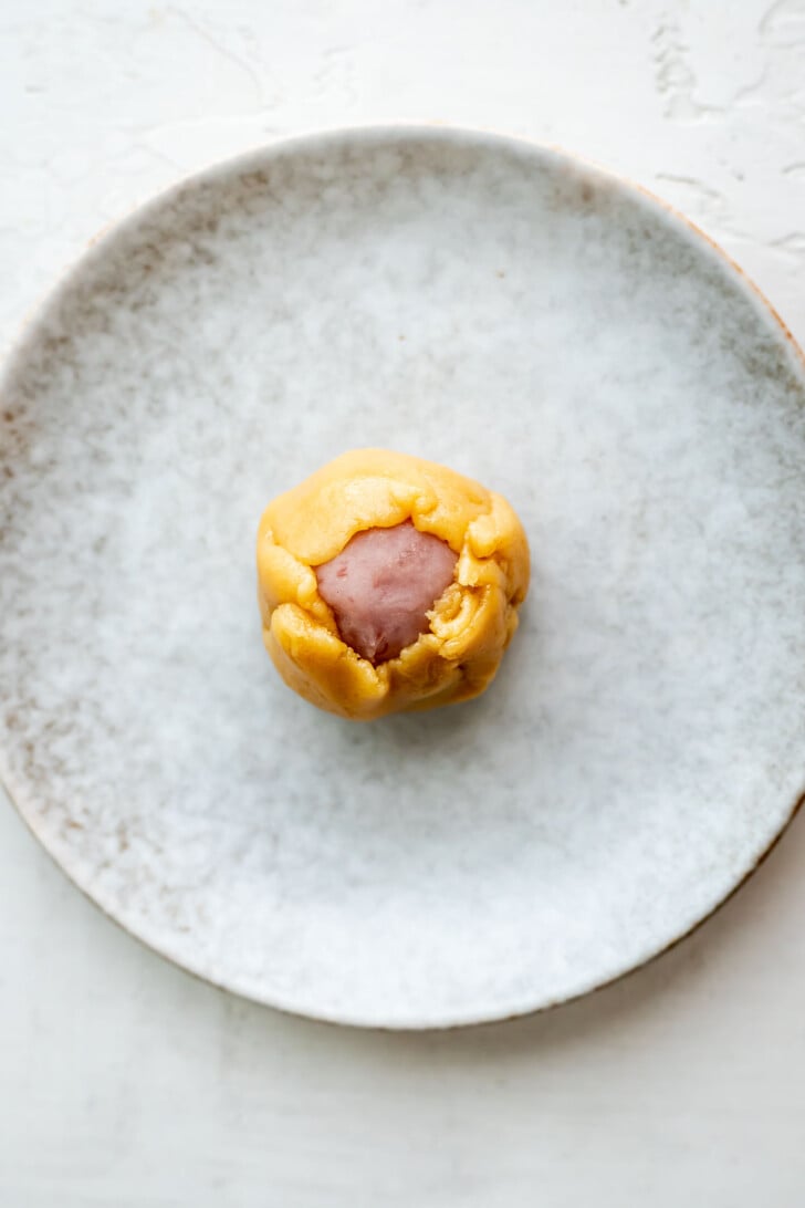 A round mooncake dough ball with a golden outer layer and a purple center sits on a white, slightly textured plate.