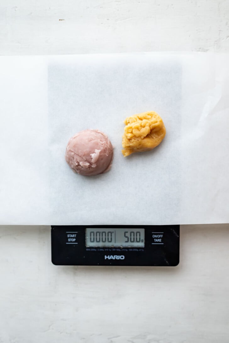 A digital scale displays 50.0 grams with a sheet of parchment paper on top. Two dough portions, one purple and one yellow, sit side by side on the paper.