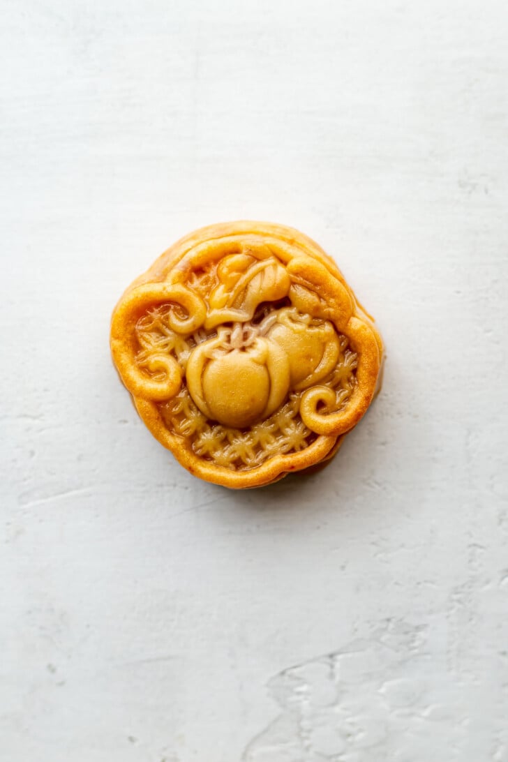 A round taro mooncake with an intricate pumpkin and vine design on top, placed on a light, textured surface.
