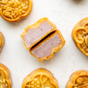 Round mooncakes with decorative tops are arranged on a white surface. One mooncake in the center is cut in half, revealing a dense, light purple taro filling inside.