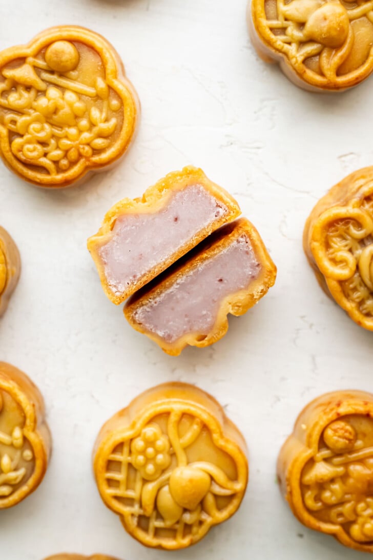 Round mooncakes with decorative tops are arranged on a white surface. One mooncake in the center is cut in half, revealing a dense, light purple taro filling inside.
