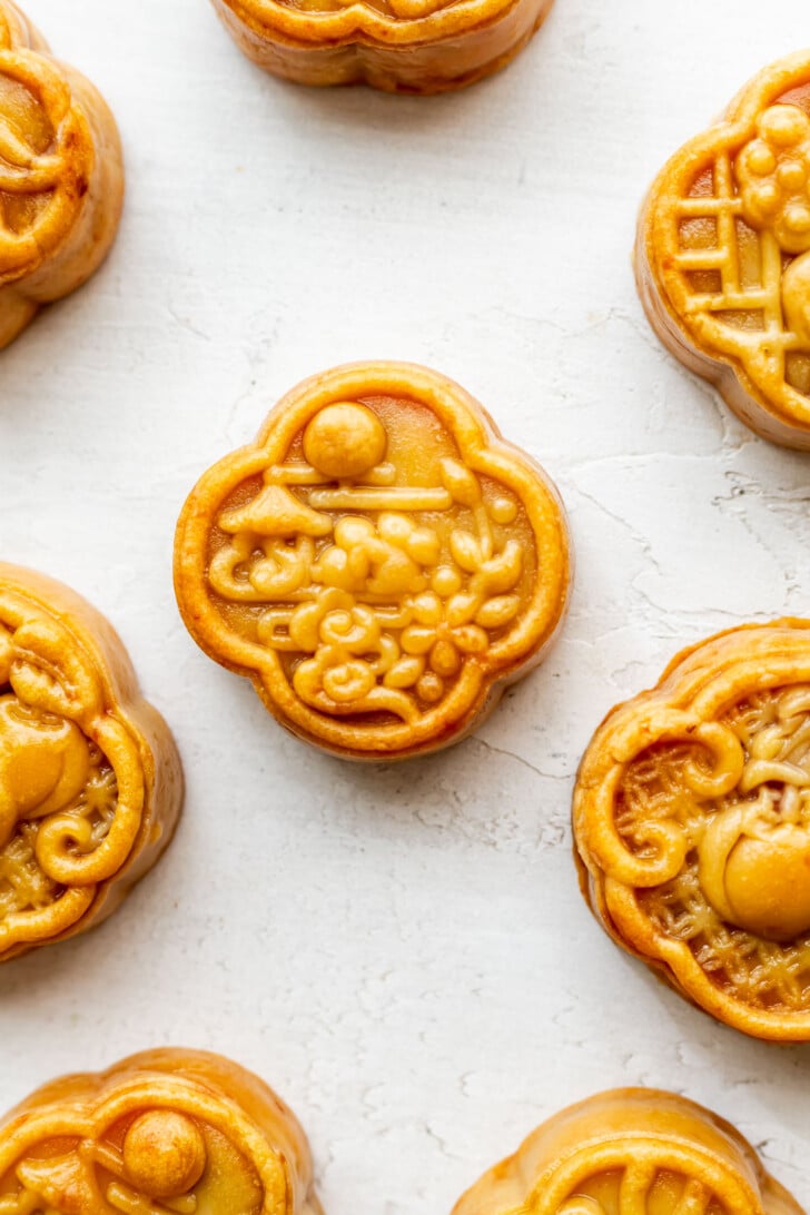 Golden mooncakes with intricate designs are arranged on a white textured surface, showcasing the elegant artistry of traditional mooncakes. One mooncake is centered, surrounded by others near the edges of the image.