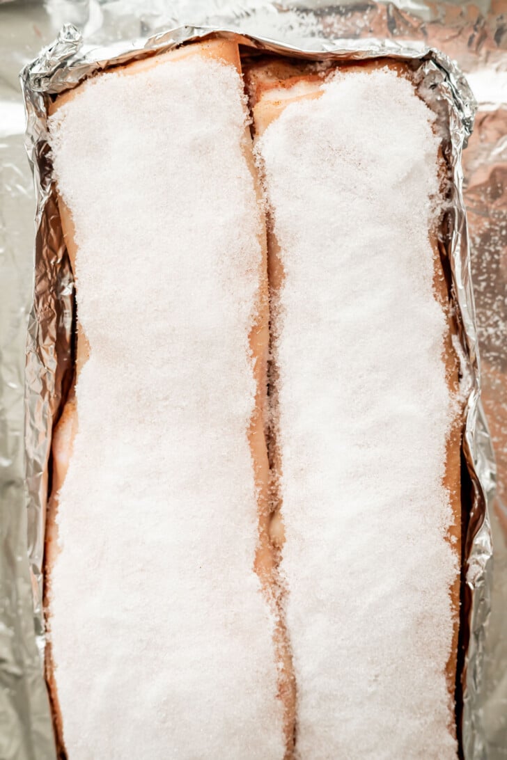 Two pork belly slabs on a foil tray are heavily coated with a thick layer of salt, ready for air frying.