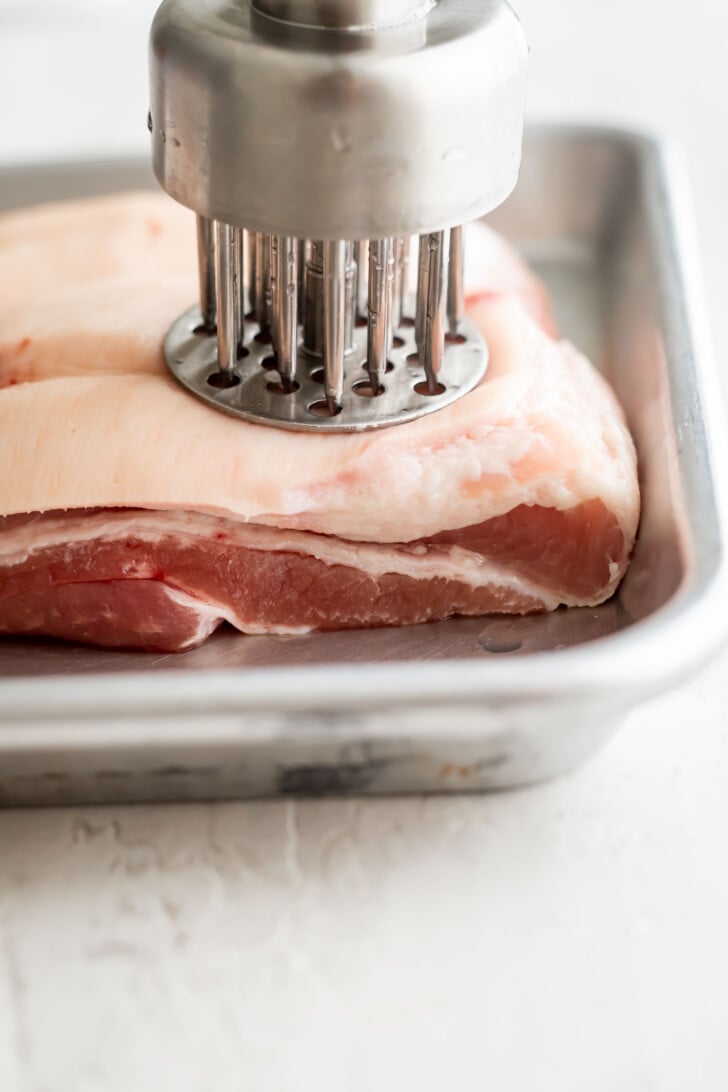 A metal meat tenderizer with multiple needles presses into a raw pork belly slab placed in a metal tray.