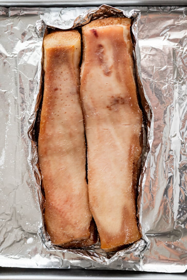 Two large pieces of pork belly after air frying for 30 minutes on a sheet of aluminum foil, set on a baking tray.