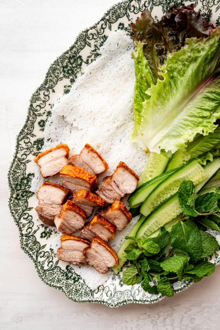 Sliced crispy pork belly, rice vermicelli, lettuce, cucumber, and mint on a decorative plate.