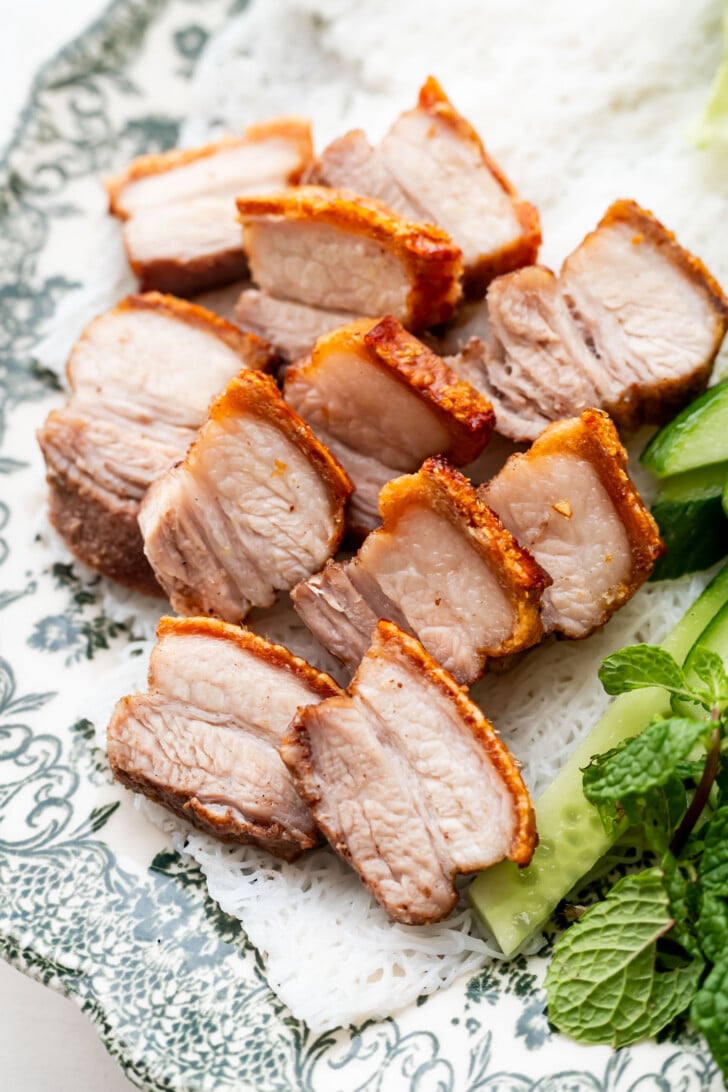 Sliced pieces of crispy pork belly arranged on a decorative plate, served with fresh cucumber and mint leaves.