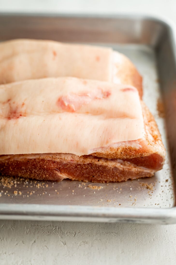 Two slabs of raw pork belly with skin on, resting in a metal baking pan. The meat is seasoned with dry spices and is ready for cooking. The surface beneath the pan is light-colored and textured.