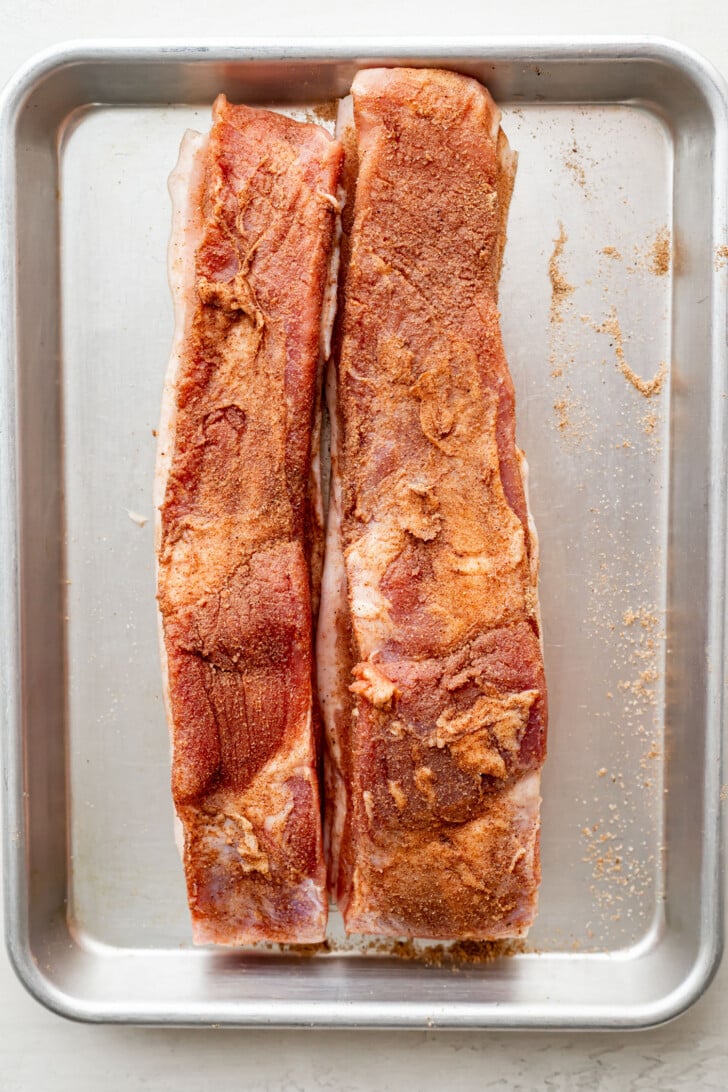 Two racks of raw pork ribs coated in dry rub seasoning sit side by side on a metal baking tray, ready for cooking.